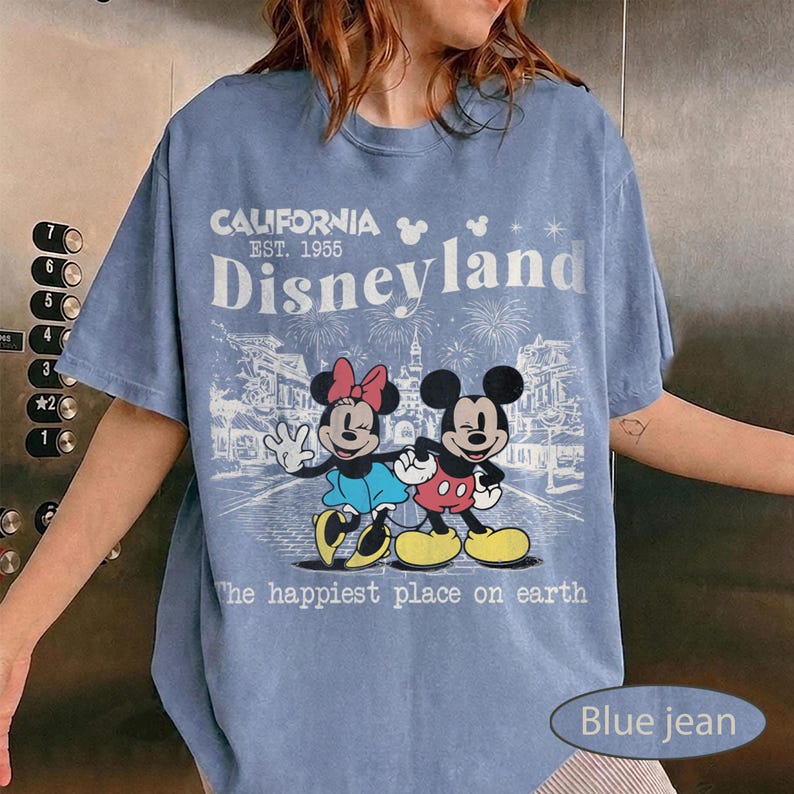May include: Blue jean t-shirt featuring a graphic of Mickey and Minnie Mouse with the text "Disneyland California Est. 1955". The design includes fireworks and the phrase "The happiest place on earth".