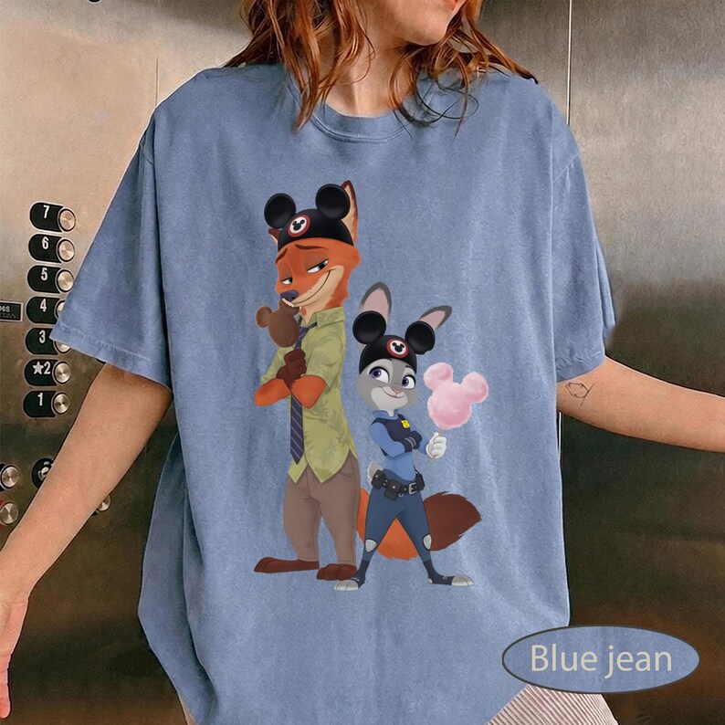 Pu&ograve; includere: T-shirt azzurra con un'illustrazione a cartoni animati di una volpe e un coniglio. La volpe indossa un cappello e una cravatta, il coniglio un'uniforme da poliziotto. Il testo "Blue jean" &egrave; in basso.