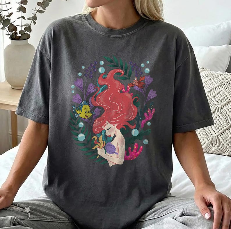 Peut inclure: T-shirt gris fonc&eacute; avec une illustration color&eacute;e d'une sir&egrave;ne aux cheveux roux, entour&eacute;e de plantes aquatiques et de cr&eacute;atures marines. Le motif comprend un poisson jaune, un crabe rouge et des bulles. Le t-shirt est de style d&eacute;contract&eacute; et confortable.