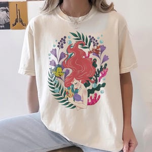 Peut inclure: T-shirt cr&egrave;me avec une illustration color&eacute;e d'une sir&egrave;ne aux cheveux roux, entour&eacute;e de plantes aquatiques et d'un poisson jaune. Le motif est centr&eacute; sur le t-shirt, cr&eacute;ant un look fantaisiste et artistique.