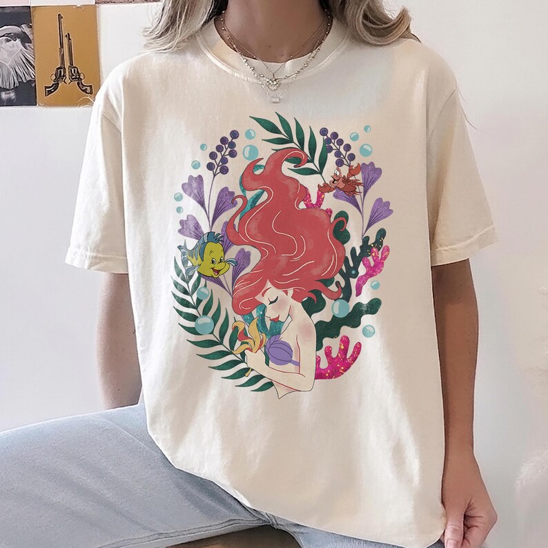 Peut inclure: T-shirt cr&egrave;me avec une illustration color&eacute;e d'une sir&egrave;ne aux cheveux roux, entour&eacute;e de plantes aquatiques et d'un poisson jaune. Le motif est centr&eacute; sur le t-shirt, cr&eacute;ant un look fantaisiste et artistique.