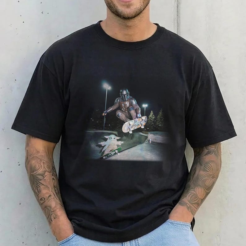 May include: A black short-sleeved crew neck t-shirt featuring a graphic print of a character in metallic armor performing a jump on a skateboard above a small green creature on a skateboard in a concrete skate park at night under bright lights.