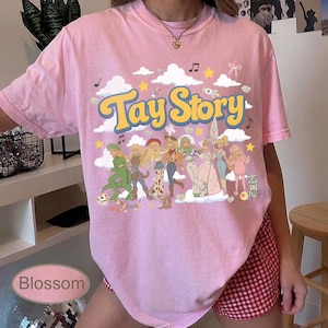 K&ouml;nnte beinhalten: Ein hellrosa T-Shirt mit kurzen &Auml;rmeln und einem Grafikdruck mit dem Text Tay Story in gelben Buchstaben. Das Design enth&auml;lt Illustrationen verschiedener Figuren in unterschiedlichen Outfits, wei&szlig;e Wolken, gelbe Sterne und Musiknoten.