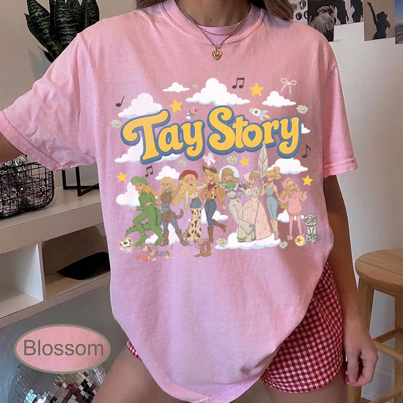 K&ouml;nnte beinhalten: Ein hellrosa T-Shirt mit kurzen &Auml;rmeln und einem Grafikdruck mit dem Text Tay Story in gelben Buchstaben. Das Design enth&auml;lt Illustrationen verschiedener Figuren in unterschiedlichen Outfits, wei&szlig;e Wolken, gelbe Sterne und Musiknoten.