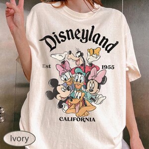 May include: Ivory-colored t-shirt featuring a graphic of Disney characters, including Mickey and Minnie Mouse, Donald and Daisy Duck, Goofy, and Pluto. The word "Disneyland" is arched across the top, with "Est 1955" and "California" below the graphic.