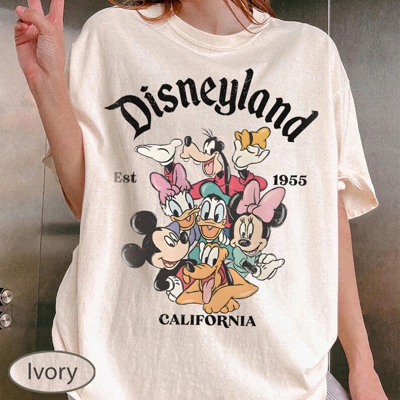 May include: Ivory-colored t-shirt featuring a graphic of Disney characters, including Mickey and Minnie Mouse, Donald and Daisy Duck, Goofy, and Pluto. The word "Disneyland" is arched across the top, with "Est 1955" and "California" below the graphic.