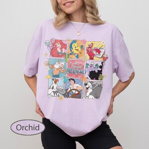 May include: Orchid-coloured t-shirt with a grid of cartoon images from The Little Mermaid. The images include Ariel, Sebastian, Flounder, and Ursula. The text "The Little Mermaid" is in the centre. The shirt is a relaxed fit.