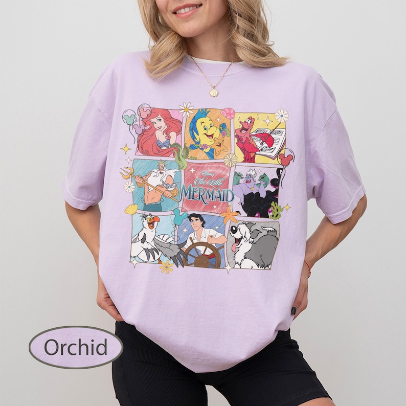 May include: Orchid-coloured t-shirt with a grid of cartoon images from The Little Mermaid. The images include Ariel, Sebastian, Flounder, and Ursula. The text "The Little Mermaid" is in the centre. The shirt is a relaxed fit.