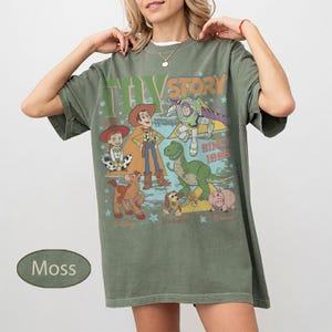 Puede incluir: Camiseta verde musgo con un dise&ntilde;o colorido de personajes de Toy Story, incluyendo a Woody y Buzz Lightyear. El texto "Toy Story" y "Since 1995" tambi&eacute;n est&aacute;n presentes. La camiseta tiene un corte holgado.