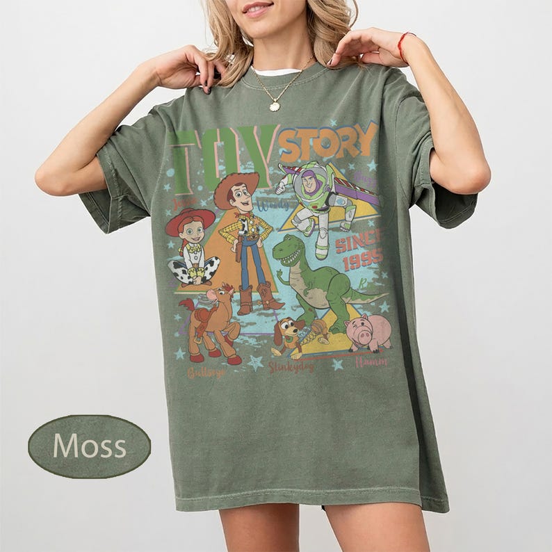 Puede incluir: Camiseta verde musgo con un dise&ntilde;o colorido de personajes de Toy Story, incluyendo a Woody y Buzz Lightyear. El texto "Toy Story" y "Since 1995" tambi&eacute;n est&aacute;n presentes. La camiseta tiene un corte holgado.