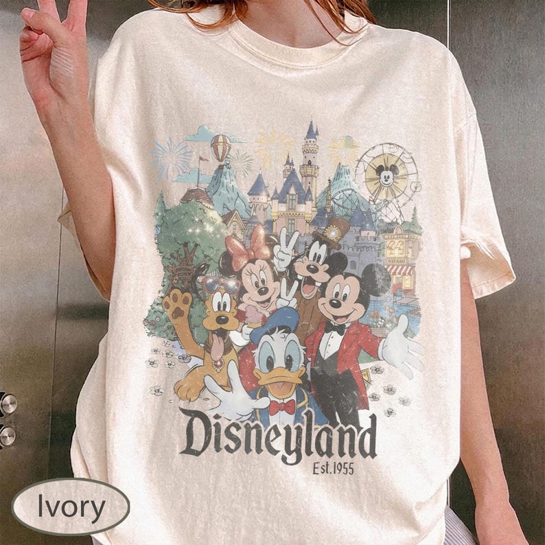 May include: An ivory short-sleeve t-shirt featuring a graphic print of Mickey Mouse, Minnie Mouse, Donald Duck, Goofy, and Pluto in front of a Disneyland castle. The text "Disneyland Est. 1955" is printed in a dark font below the character illustration.