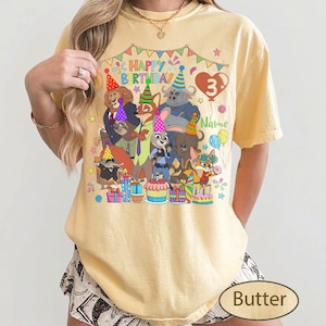 May include: Butter-colored t-shirt featuring a colorful graphic of cartoon animals, party hats, and the text "Happy Birthday 3". The design includes a heart-shaped balloon and the word "Name".