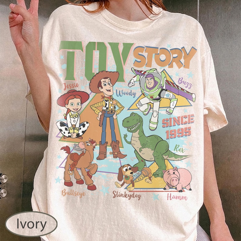 Puede incluir: Camiseta color marfil con un estampado de los personajes de Toy Story, incluyendo a Woody, Buzz, Jessie y Rex. El texto "TOY STORY" est&aacute; en la parte superior, con "SINCE 1995" debajo. La camiseta tiene cuello redondo y mangas cortas.