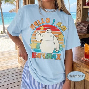 May include: Light blue t-shirt featuring a graphic of Baymax from Big Hero 6. The design includes the text "Hello I Am Baymax" in a colorful, retro style, with stars and a circular background.