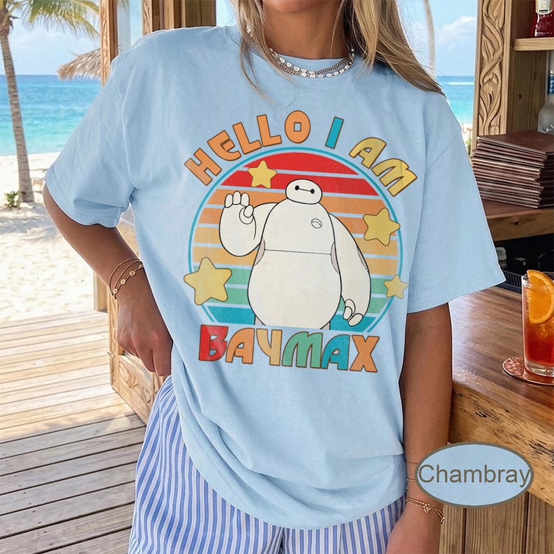 May include: Light blue t-shirt featuring a graphic of Baymax from Big Hero 6. The design includes the text "Hello I Am Baymax" in a colorful, retro style, with stars and a circular background.