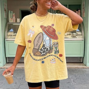 May include: A pale yellow short-sleeve t-shirt featuring a graphic print of a cowboy boot with a cow-print pattern, a red hat, and blocks labeled T S. The text TAY STORY is printed on the boot. Additional graphics include a bow, flowers, and a beaded string.