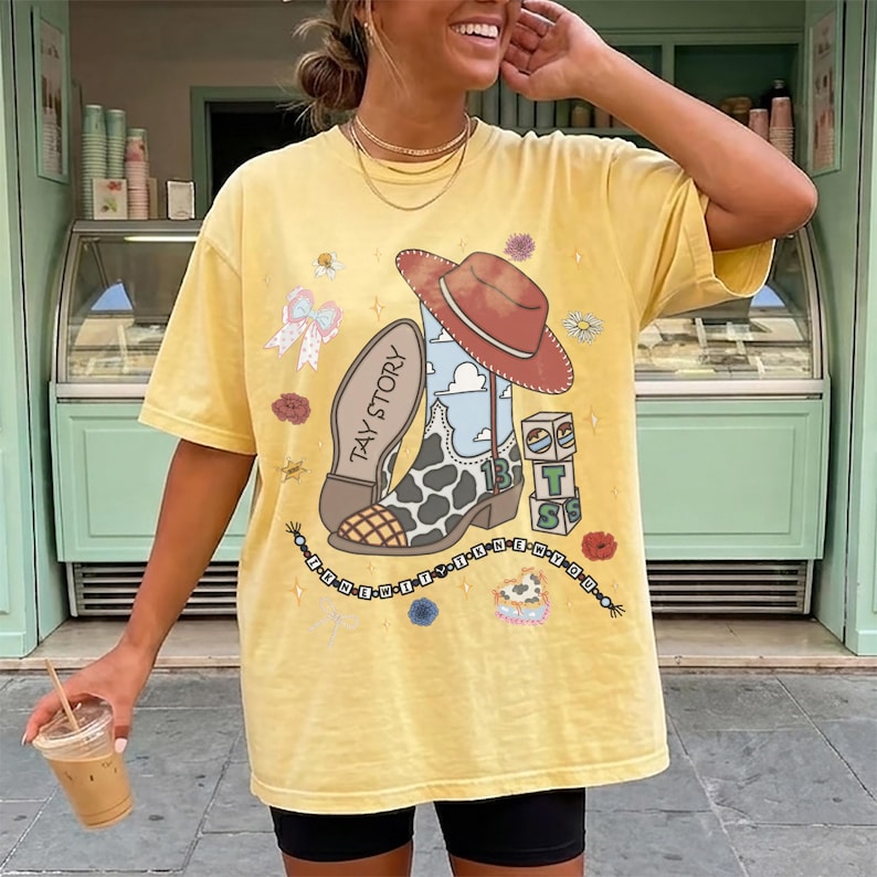 May include: A pale yellow short-sleeve t-shirt featuring a graphic print of a cowboy boot with a cow-print pattern, a red hat, and blocks labeled T S. The text TAY STORY is printed on the boot. Additional graphics include a bow, flowers, and a beaded string.