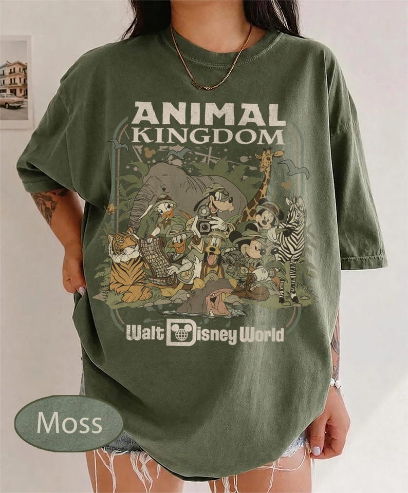 May include: Moss green t-shirt with a graphic featuring the words "ANIMAL KINGDOM" and "Walt Disney World." The graphic includes cartoon animals like an elephant, giraffe, and zebra, along with Disney characters.
