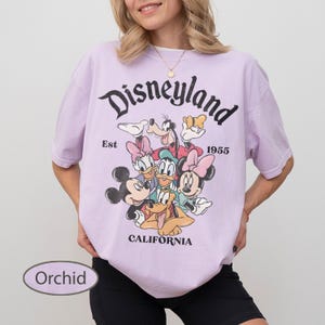 May include: Orchid-colored t-shirt featuring a Disneyland graphic with Mickey Mouse, Minnie Mouse, Donald Duck, Goofy, and other Disney characters. The shirt has the text "Disneyland" above the characters, with "Est 1955" and "California" below.