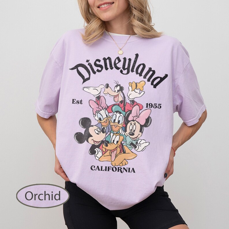 May include: Orchid-colored t-shirt featuring a Disneyland graphic with Mickey Mouse, Minnie Mouse, Donald Duck, Goofy, and other Disney characters. The shirt has the text "Disneyland" above the characters, with "Est 1955" and "California" below.