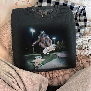 May include: A charcoal gray crewneck sweatshirt featuring a graphic print of a Mandalorian character performing a skateboard trick in a bowl, with a small Grogu character on a separate skateboard below, set against a dark night background.