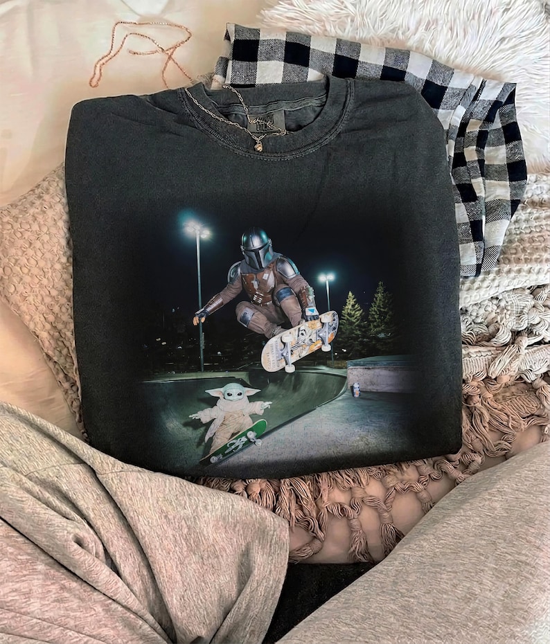 May include: A charcoal gray crewneck sweatshirt featuring a graphic print of a Mandalorian character performing a skateboard trick in a bowl, with a small Grogu character on a separate skateboard below, set against a dark night background.