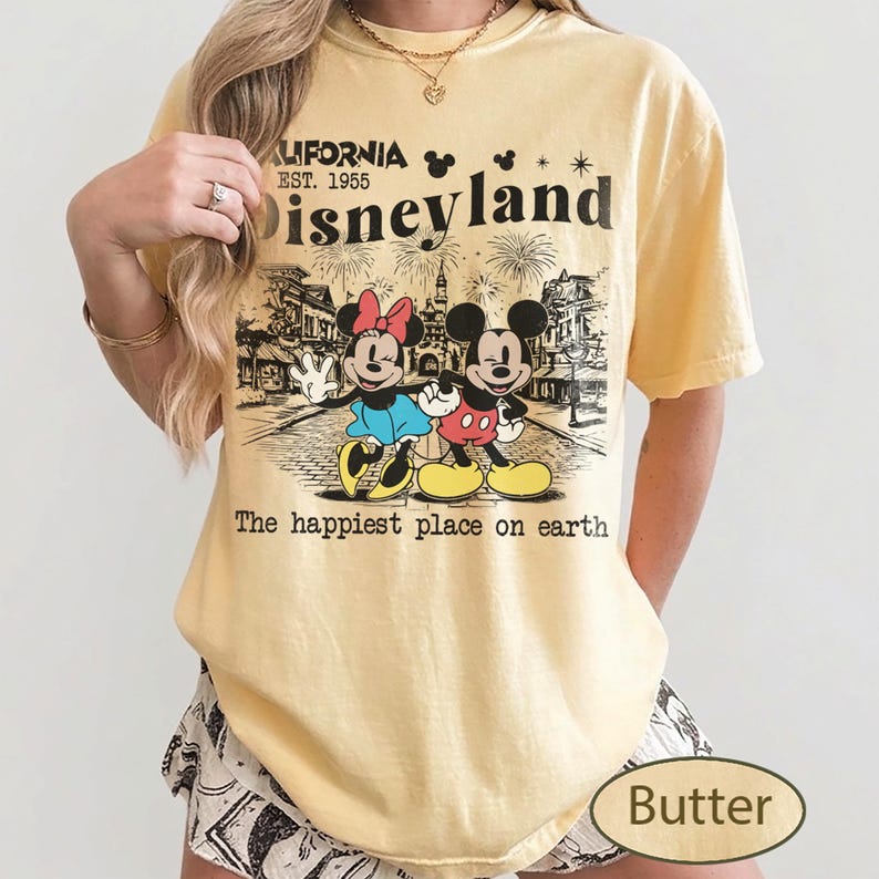 May include: Butter-colored t-shirt featuring a graphic of Mickey and Minnie Mouse in front of Disneyland. The shirt includes the text "California Est. 1955 Disneyland" and "The happiest place on earth."