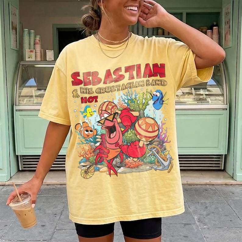 以下が含まれることがあります： SEBASTIAN & HIS CRUSTACEAN BAND HOTというテキストがプリントされた、ペールイエローの半袖Tシャツ。サングラスをかけてマラカスを持った赤いカニが、色鮮やかなサンゴや魚たちに囲まれているデザインが描かれています。