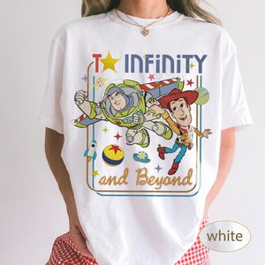 May include: White t-shirt featuring a colorful graphic of Buzz Lightyear and Woody from Toy Story, with the text "To Infinity and Beyond." The design includes a star, rocket, and other playful elements.