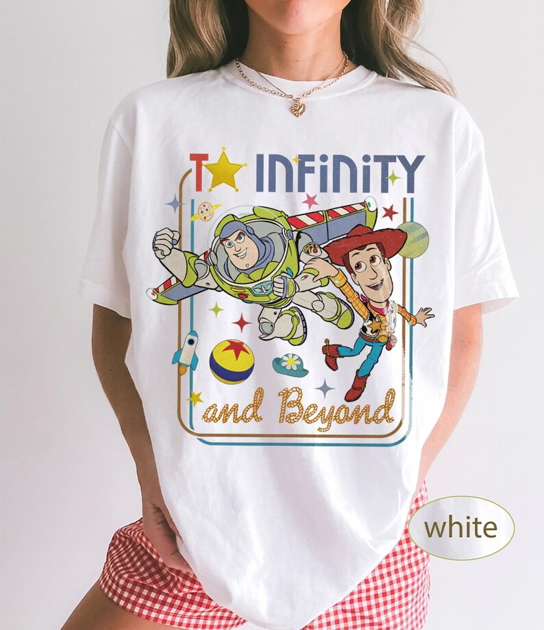 May include: White t-shirt featuring a colorful graphic of Buzz Lightyear and Woody from Toy Story, with the text "To Infinity and Beyond." The design includes a star, rocket, and other playful elements.