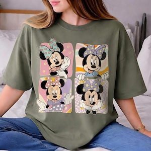 May include: Olive green t-shirt featuring a four-panel design of Minnie Mouse. Each panel showcases a different pose of Minnie, with polka dot bows and colorful backgrounds. The shirt is a casual, oversized fit.
