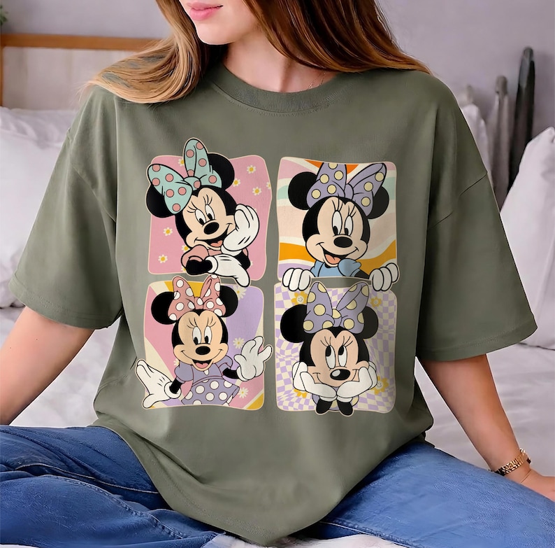 May include: Olive green t-shirt featuring a four-panel design of Minnie Mouse. Each panel showcases a different pose of Minnie, with polka dot bows and colorful backgrounds. The shirt is a casual, oversized fit.