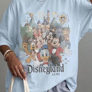 May include: A light blue t-shirt featuring a graphic print of Mickey Mouse, Minnie Mouse, Donald Duck, Goofy, and Pluto in front of a castle and ferris wheel. The text "Disneyland Est. 1955" is printed in a dark font below the characters.