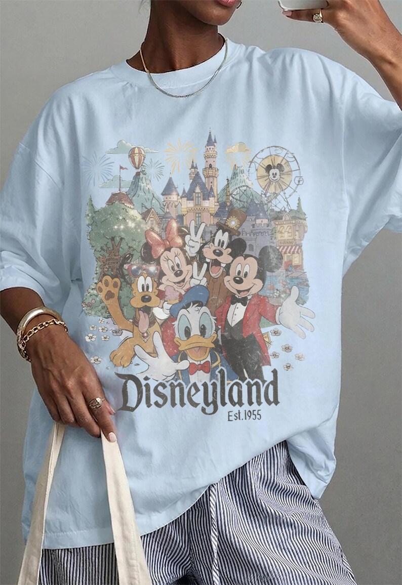 May include: A light blue t-shirt featuring a graphic print of Mickey Mouse, Minnie Mouse, Donald Duck, Goofy, and Pluto in front of a castle and ferris wheel. The text "Disneyland Est. 1955" is printed in a dark font below the characters.