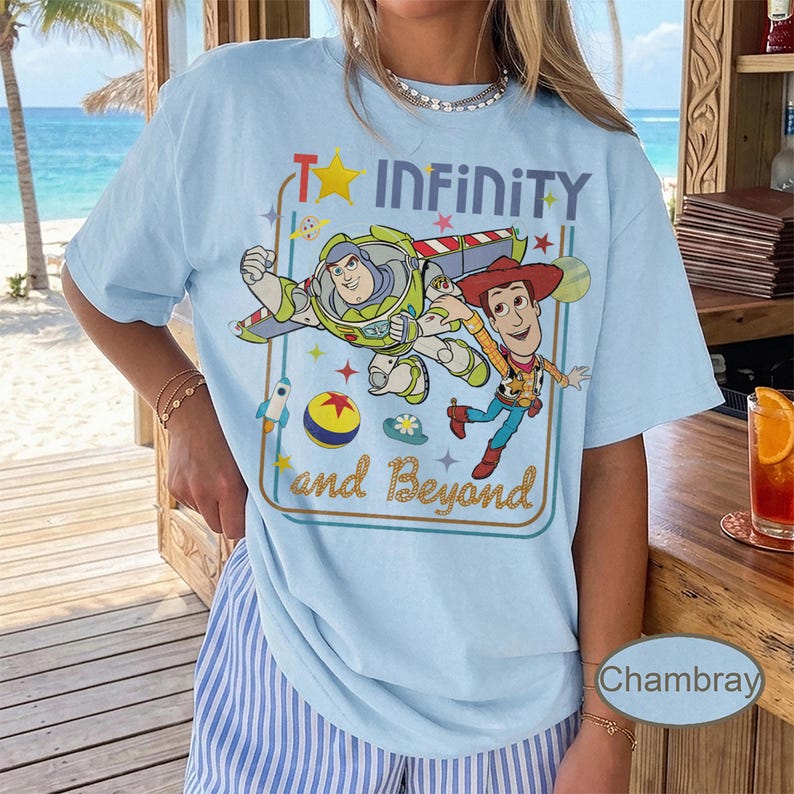 May include: Light blue t-shirt featuring Buzz Lightyear and Woody from Toy Story, with the text "To Infinity and Beyond." The design includes a rocket, a ball, and stars. The shirt is made of a soft material.