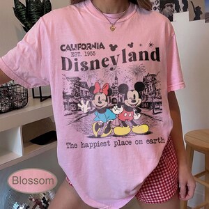 May include: Pink t-shirt with a Disneyland graphic featuring Mickey and Minnie Mouse. The design includes the text "CALIFORNIA EST. 1955 Disneyland" and "The happiest place on earth."