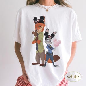 Pu&ograve; includere: T-shirt bianca con un'illustrazione a cartoni animati di Nick Wilde e Judy Hopps di Zootropolis, con orecchie di Topolino. Nick tiene in mano un piccolo peluche e Judy tiene in mano zucchero filato rosa. La parola "bianco" &egrave; in un ovale bianco.
