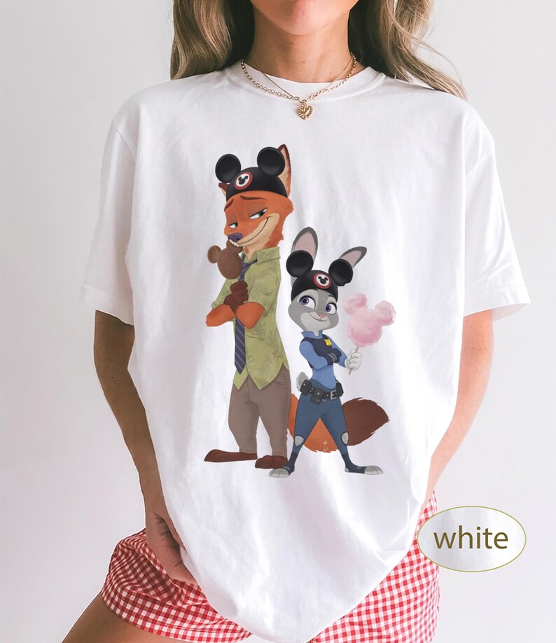 Pu&ograve; includere: T-shirt bianca con un'illustrazione a cartoni animati di Nick Wilde e Judy Hopps di Zootropolis, con orecchie di Topolino. Nick tiene in mano un piccolo peluche e Judy tiene in mano zucchero filato rosa. La parola "bianco" &egrave; in un ovale bianco.
