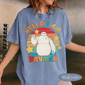 May include: Blue jean t-shirt featuring a graphic of Baymax from Big Hero 6. The design includes the text "HELLO I AM BAYMAX" in orange and red, with a circular background of colorful stripes and yellow stars.