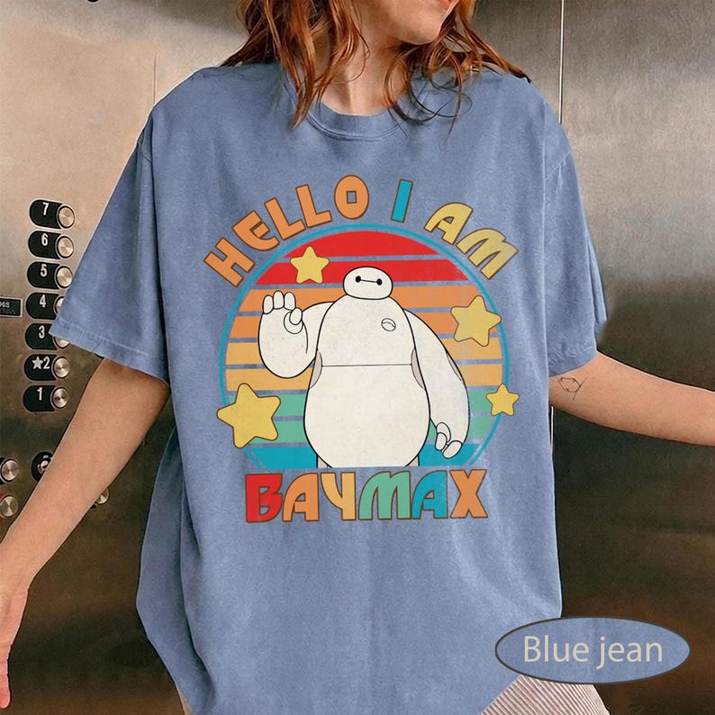 May include: Blue jean t-shirt featuring a graphic of Baymax from Big Hero 6. The design includes the text "HELLO I AM BAYMAX" in orange and red, with a circular background of colorful stripes and yellow stars.
