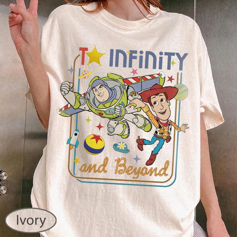 May include: Ivory-colored t-shirt featuring a graphic of Buzz Lightyear and Woody from Toy Story. The design includes the text "To Infinity and Beyond" with stars and other playful elements. The shirt has a crew neck and short sleeves.