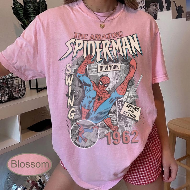 May include: Pink t-shirt featuring a graphic of Spider-Man in action, with the text "The Amazing Spider-Man" and "New York." The design includes the year "1962" and the words "Spring into Action."