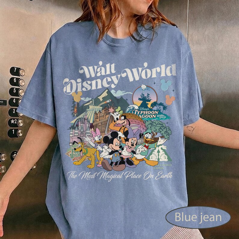 May include: Blue jean colored t-shirt featuring a colorful graphic of Disney characters and the text "Walt Disney World". The design includes Mickey Mouse, Minnie Mouse, Donald Duck, and Goofy. The shirt also has the text "The Most Magical Place On Earth".