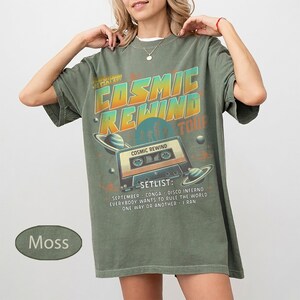 K&ouml;nnte beinhalten: Moosgr&uuml;nes T-Shirt mit Retro-Grafikdesign. Das Design zeigt den Text "COSMIC REWIND TOUR" mit einem Kassettenrekorder und Planeten. Die Setlist enth&auml;lt Songs wie "September" und "One Way or Another".