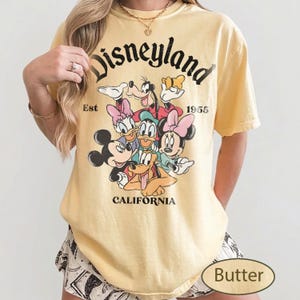 May include: Butter-colored t-shirt featuring the word "Disneyland" in a black script, with the text "Est 1955" and "California" below. The shirt displays a graphic of Mickey Mouse, Minnie Mouse, Donald Duck, Daisy Duck, Goofy, and Pluto.