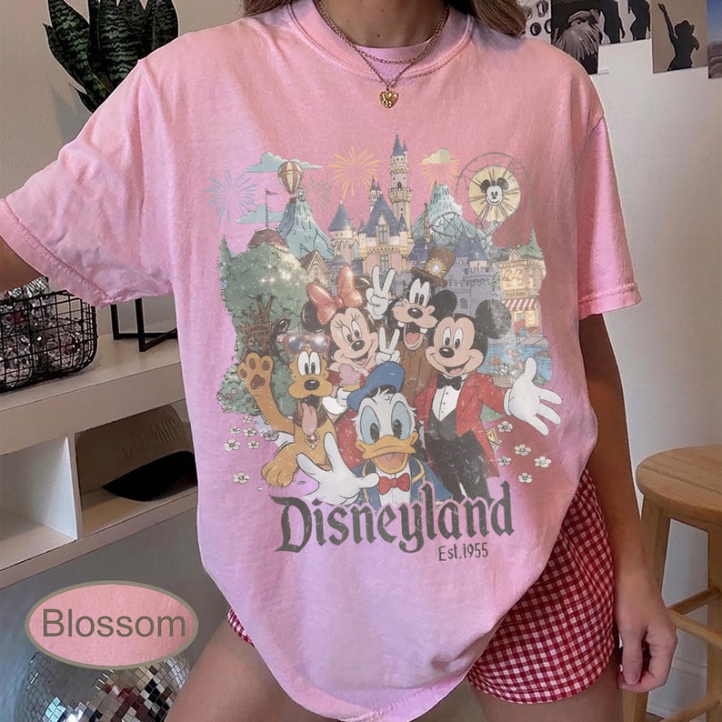 May include: A light pink short-sleeve t-shirt featuring a graphic print of Mickey Mouse, Minnie Mouse, Donald Duck, and Pluto in front of a castle. The text reads Disneyland Est. 1955. The shirt has a crew neckline and a relaxed fit design.