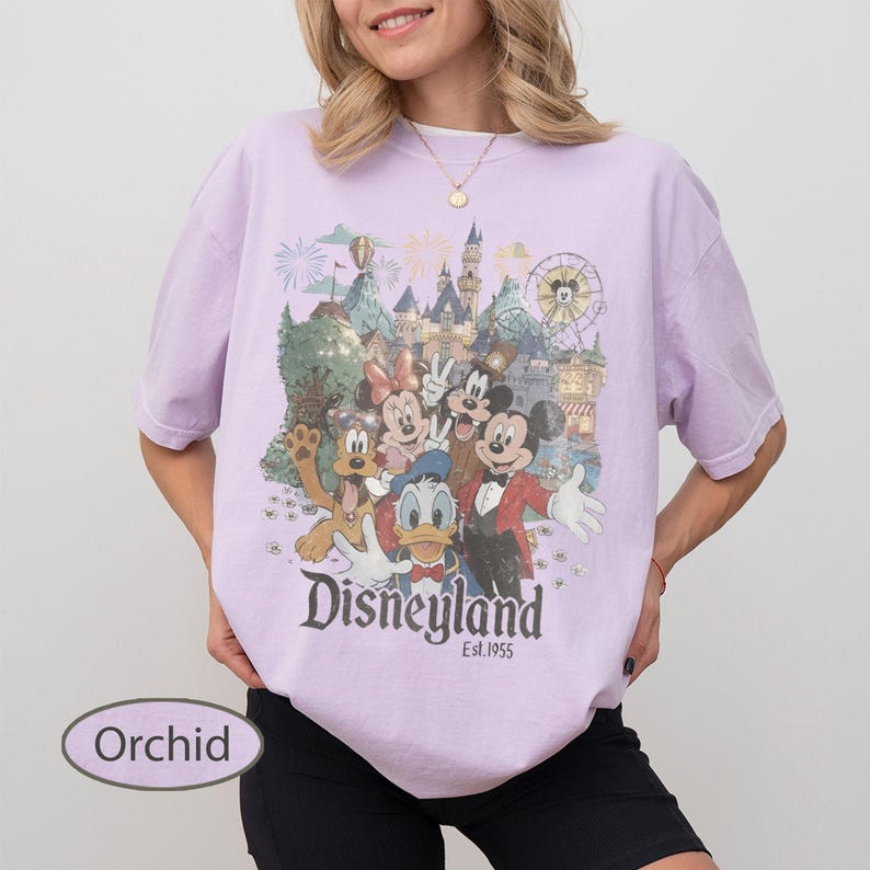 May include: A light purple short-sleeve t-shirt featuring a graphic print of Mickey Mouse, Minnie Mouse, Donald Duck, Goofy, and Pluto in front of a castle. The text Disneyland Est. 1955 is printed below the characters. The word Orchid is visible.