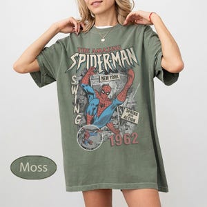 May include: Moss green t-shirt featuring a vintage-style graphic of Spider-Man in action. The design includes the text "THE AMAZING SPIDER-MAN NEW YORK" and "1962". The shirt has short sleeves and a relaxed fit.