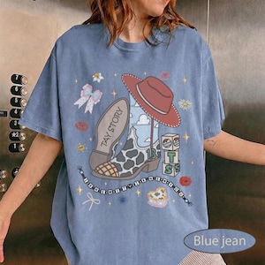 May include: A blue jean colored t-shirt featuring a graphic print of a cow print cowboy boot, a red cowboy hat, building blocks with the letters T, S, and the number 13, a bow, and text reading TAY STORY and I KNEW IT I KNEW YOU.