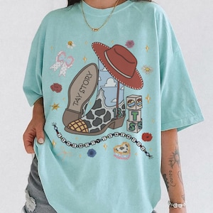 May include: A light blue t-shirt featuring a graphic print of a cowboy boot with a cow print pattern, a red cowboy hat, building blocks with the letters T, S, and the number 13, and a beaded necklace graphic with the text I KNEW IT I KNEW YOU.